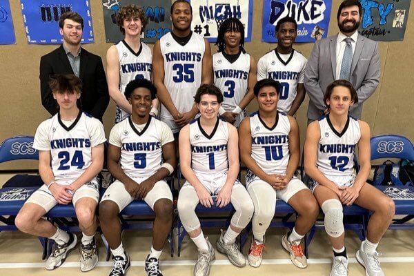 A high school basketball team poses for a group photo in uniforms with coaches, sitting and standing in two rows indoors.