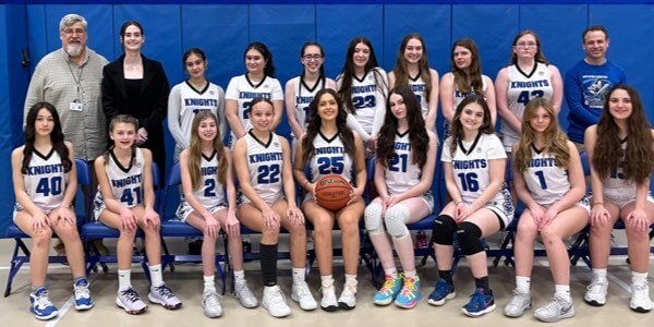Group photo of a girls' basketball team in uniforms, seated and standing in two rows, with two adults at each end. One player in the center holds a basketball.