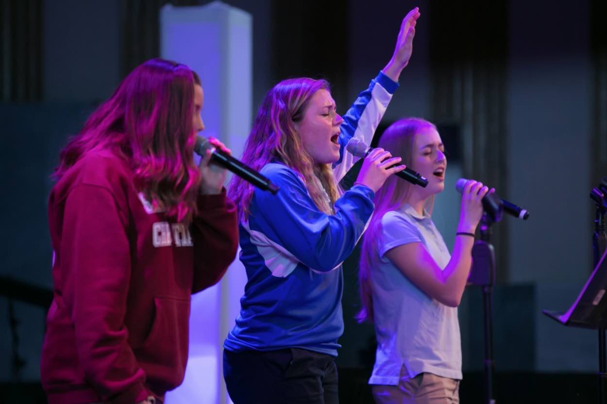 Three young women sing into microphones on a stage, with one raising her hand while performing under purple and blue lighting.
