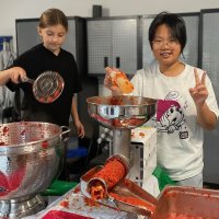 Two people process tomatoes using a food mill and metal strainer in a kitchen; one holds a strainer, the other flashes a peace sign while holding tomato pulp.