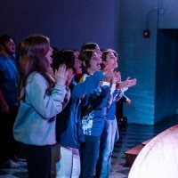 A group of people standing indoors, clapping and facing a stage or platform, with blue lighting illuminating the scene.