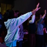 A group of people stand indoors with their arms raised, appearing to participate in a group activity or event.