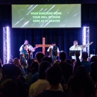 A band performs on stage at a church service with a wooden cross in the background and lyrics projected on a screen above.