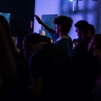 A group of people standing in a dimly lit room; one person in the middle raises their hand.