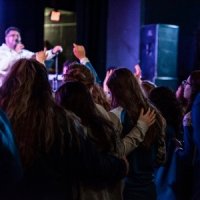 A crowd stands closely together facing a performer on stage. The performer is pointing toward the audience under stage lighting.