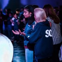 A group of people stand in a dimly lit room, some with hands raised or open, appearing to participate in a communal or reflective activity.