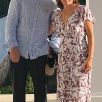 Two people standing side by side outdoors, one wearing a light blue shirt and dark pants, the other in a white and purple floral dress, both smiling at the camera.