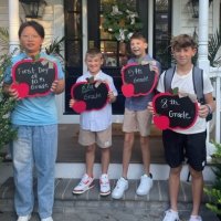 Four boys stand on a porch holding apple-shaped signs indicating their first day of 5th, 6th, 7th, and 8th grades.