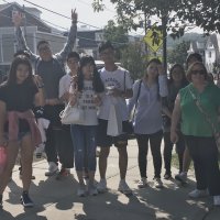 A group of people stand together on a sidewalk in a residential neighborhood on a sunny day, some smiling and posing for the photo.