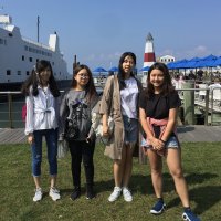 Four young women stand on grass near a docked ferry and a red-and-white lighthouse, with blue umbrellas visible in the background on a sunny day.