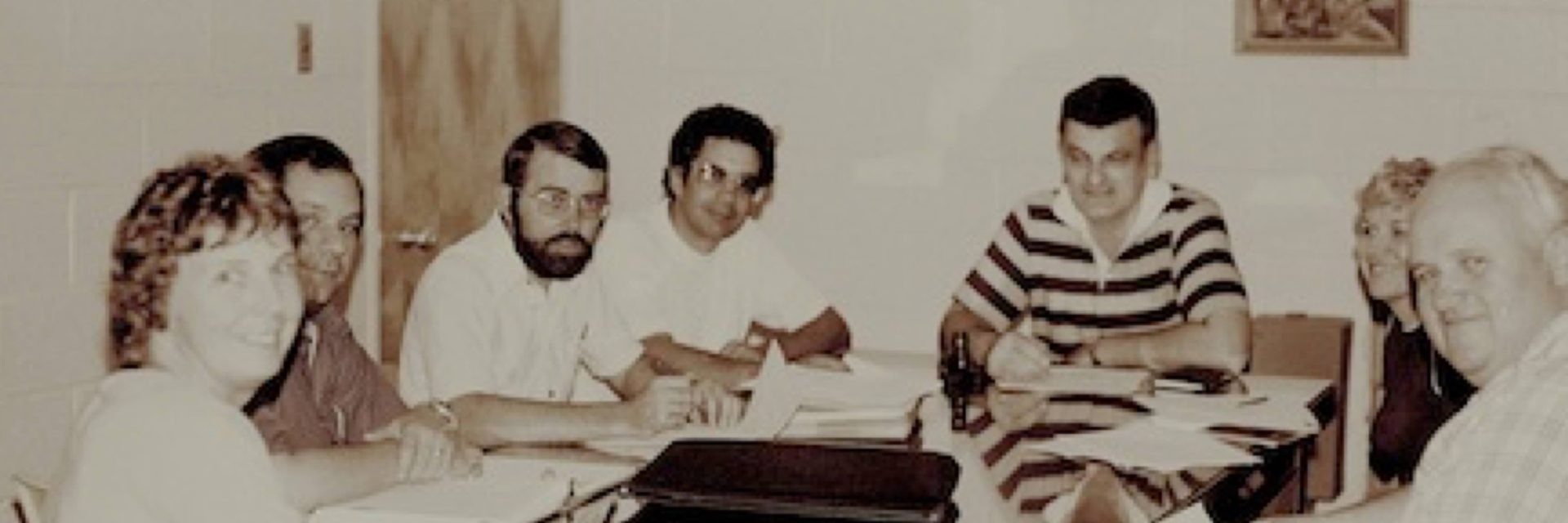Seven people sit around a table in a meeting room, engaged in discussion with papers and folders in front of them.