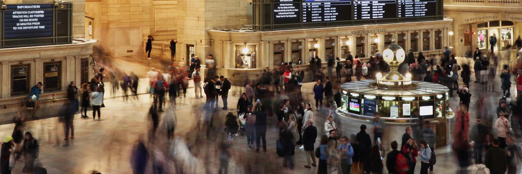A busy hall at Grand Central Terminal with people walking and gathering near the iconic clock and information booths. Departure boards are visible in the background.