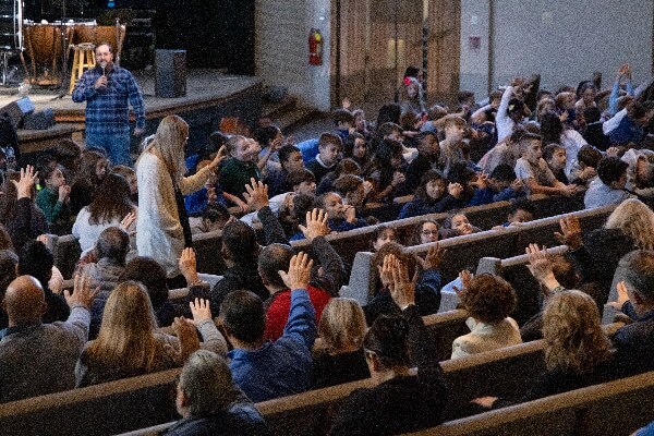 A large audience sits in rows inside an auditorium as many people raise their hands; a man and a woman stand among them, interacting with the crowd.