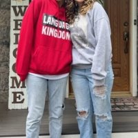 Two people stand side by side on a porch in front of a door. One wears a red hoodie and jeans, the other a gray sweatshirt and ripped jeans. A