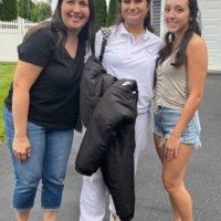 Three women stand together outdoors on a driveway, smiling at the camera. One woman is holding a black jacket, and all are dressed casually.