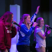 Three young women sing into microphones on stage; one has her arm raised. The lighting is purple and blue.