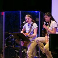 Two students in uniform perform on stage; one plays guitar and sings into a microphone, the other sits beside her, also singing, with drums visible in the background.