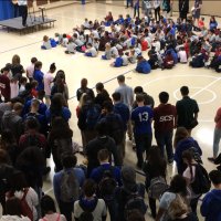 A large group of students gather in a gymnasium, with some seated on the floor in front of a stage and others standing at the back, facing forward.