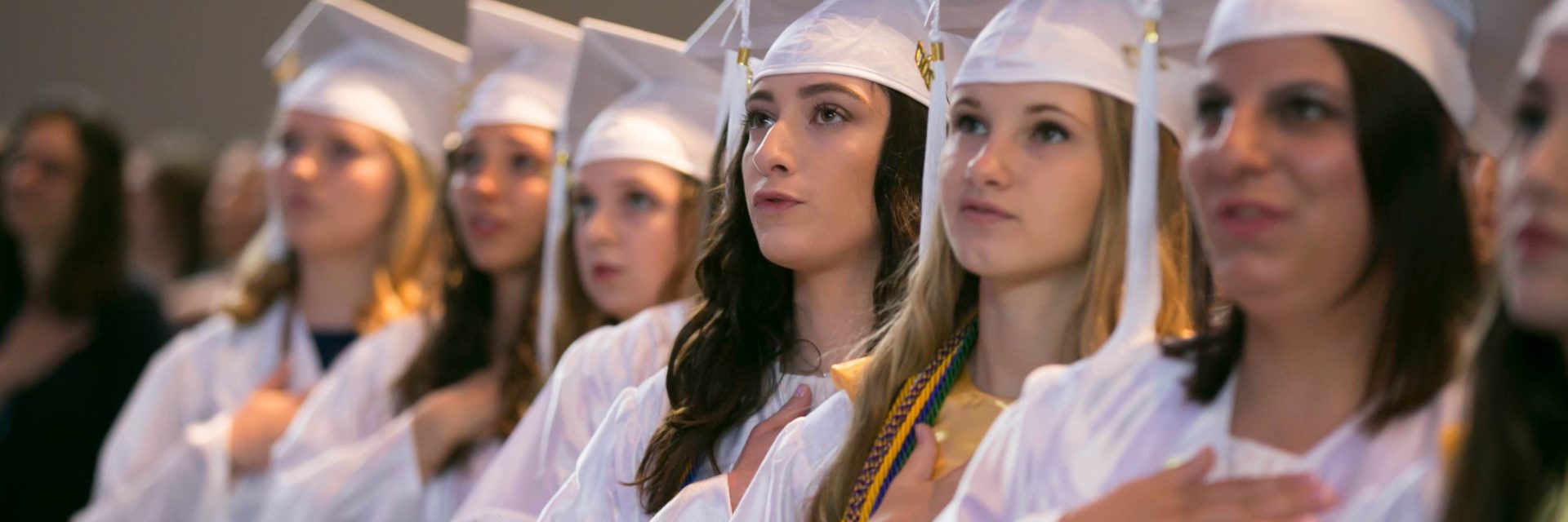 A row of graduates in white caps and gowns stand with hands over their hearts, participating in a ceremony.