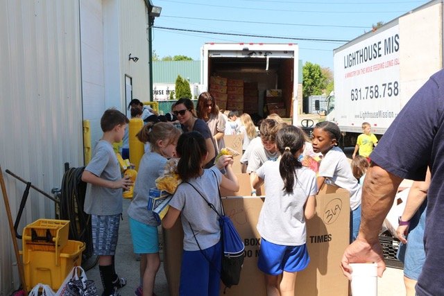 Children and adults gather around boxes, sorting items near a truck outside a building labeled