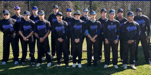 A group of sixteen baseball players and two coaches stand in uniform in front of a chain-link fence on a sunny day.