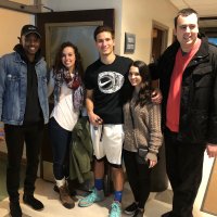 Five people stand together indoors, posing for a photo and smiling at the camera. They are dressed casually and appear to be in a hallway or common area.