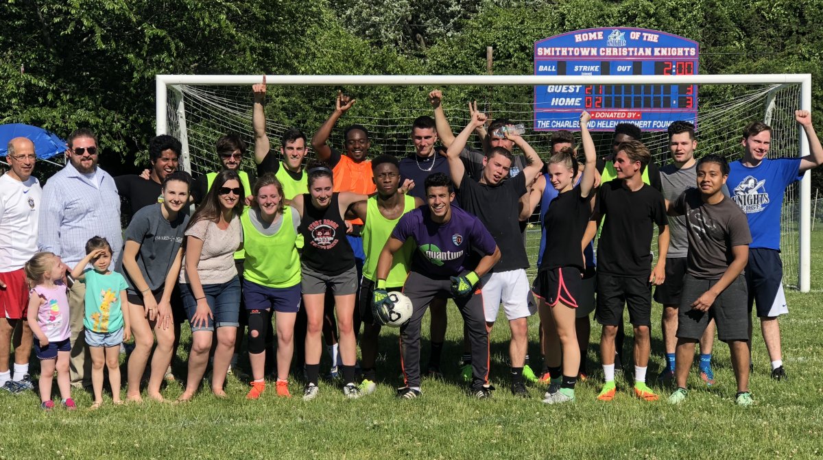 A group of men, women, and children pose in front of a soccer goal and scoreboard on a sunny day, with some holding a soccer ball and others raising their arms.