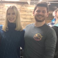 Two people are standing next to each other indoors, smiling at the camera. The background features wood paneling.