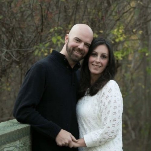 A man and woman stand close together outdoors by a wooden railing, smiling at the camera, with trees in the background.