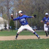 A baseball player in a blue uniform prepares to throw the ball on a field, while another player runs in the background.