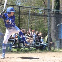 A baseball player in a blue uniform prepares to swing at bat, with spectators watching from metal bleachers behind a chain-link fence.