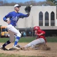 A baseball player in blue prepares to catch a ball as a player in red slides into a base, kicking up dust on a baseball field.